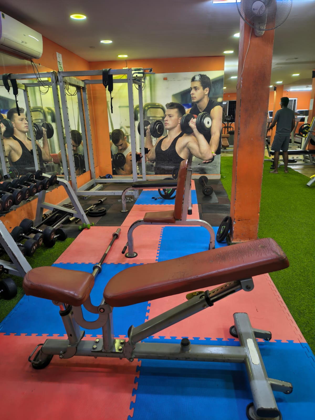 Adjustable benches on foam mats beside a dumbbell rack, power rack and wall mirrors