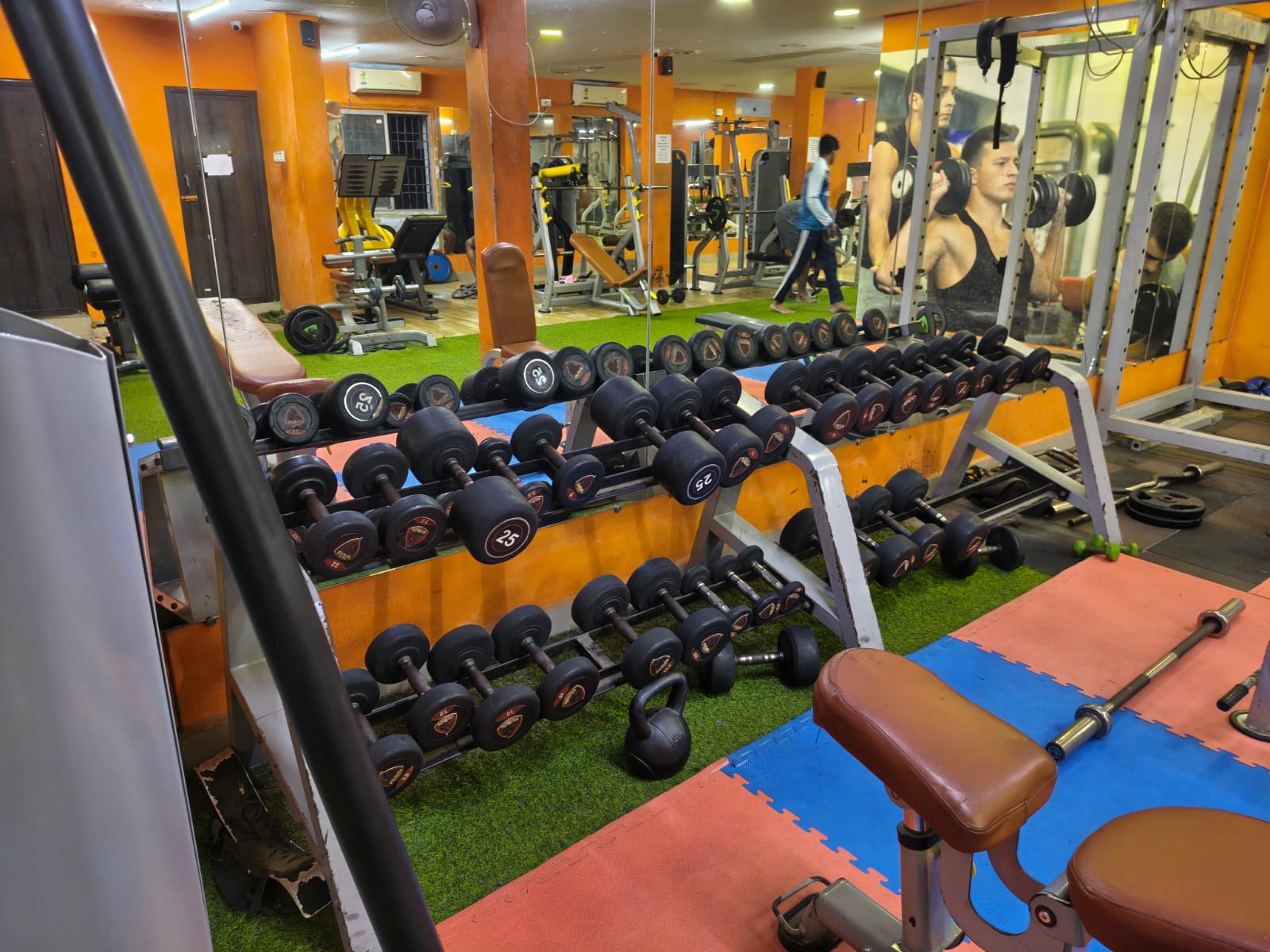 Two-tier racks of rubber dumbbells and a kettlebell on turf, beside a mirrored wall and power rack