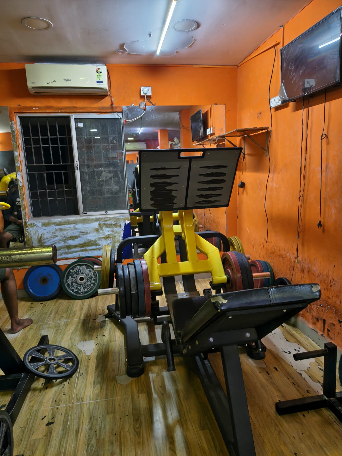 Plate-loaded leg press stacked with bumper plates, with a wall-mounted TV and air conditioner above